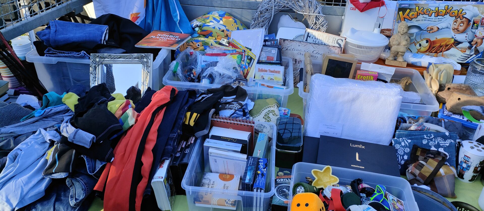 Prall gefüllter Verkaufsstand auf einem Flohmarkt: gebrauchte Kleidung, Bücher, Spielzeug, Geschirr und Deko in Sortierkisten auf einer Decke, im Morgenlicht