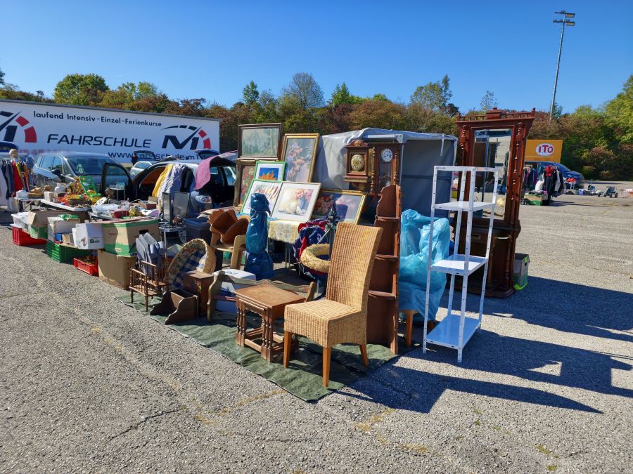 Flohmarktstand mit Holzmöbeln auf Asphalt: ein Rattanstuhl, ein Satztisch, ein hoher dunkler Garderobenschrank mit Spiegel, alte Wanduhren und mehrere gerahmte Bilder auf einer grünen Plane. Dahinter Autos mit geöffneten Kofferräumen und ein weißer Lkw.