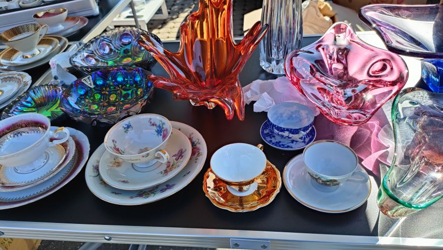 Farbige Kunstglas-Schalen und -Vasen neben mehreren dekorativen Sammeltassen mit Untertassen auf einem schwarzen Flohmarkt-Tisch.