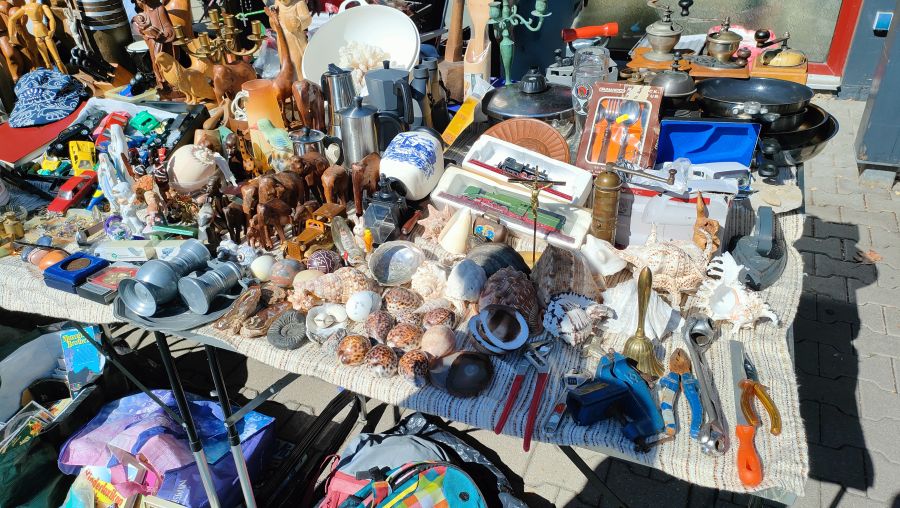Trödeltisch mit vielen Muscheln und Meeresschnecken, geschnitzten Holztieren, Zinnbechern, Modelldampfloks und Werkzeug auf einem Flohmarkt.