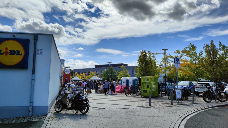 Überblick über den Flohmarkt am Rudeltplatz in Erlangen: Links das Lidl-Gebäude mit Logo, in der Bildmitte weiße Faltpavillons mit Ständen und Besucherinnen und Besuchern, rechts zwei blaue mobile Toilettenkabinen, ein grüner Altkleider-Automat, Fahrradbügel mit E-Bikes und geparkte Autos sowie ein Aufsteller mit den Marktzeiten. Gepflasterter Platz, sonniger Tag mit Wolken.