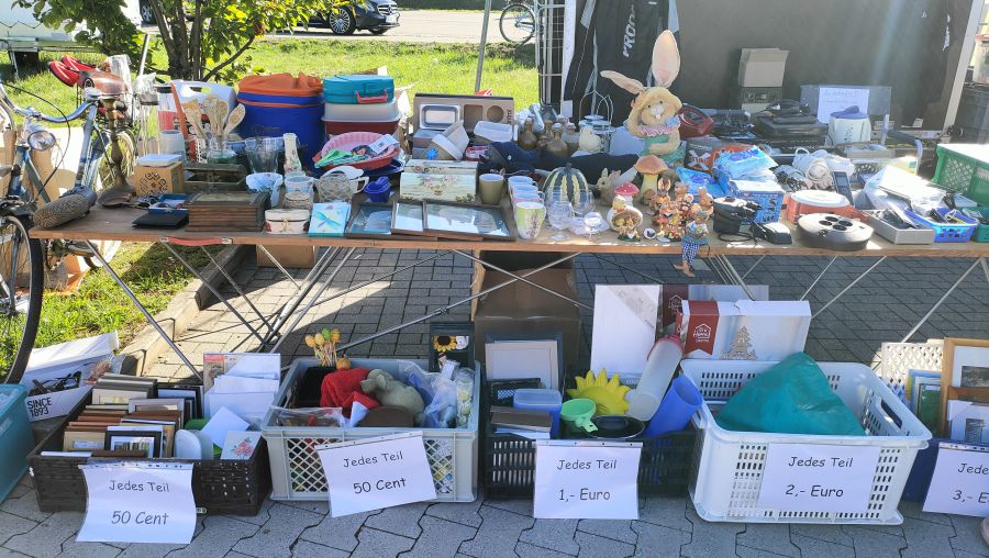 Flohmarktstand mit Haushalts-Kleinkram auf dem Tisch und mehreren Sortierkisten mit 50-Cent- bis 3-Euro-Preisschildern darunter.