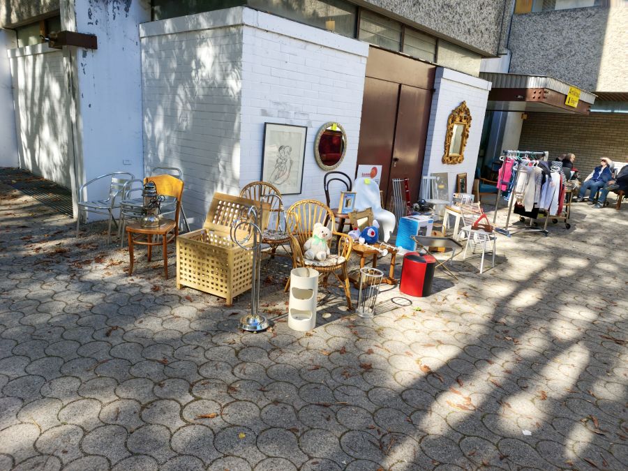 Möbel und Wohnaccessoires vor einer weißen Backsteinwand: Aluminiumstühle, Bugholzstuhl mit Sturmlaterne, Rattansessel mit Teddybär, Holztruhe, Schirmständer, roter Pouf und Spiegel mit goldenem Rahmen. Rechts steht ein Kleiderständer mit Damenmode auf dem gepflasterten Boden.