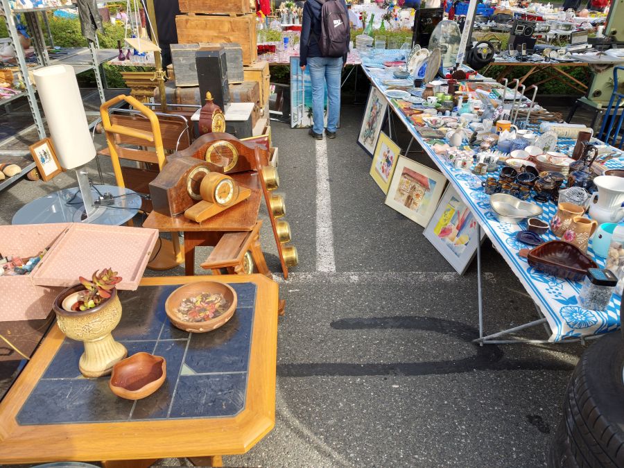 Blick durch eine Marktgasse mit Antiktischen voller Uhren, Koffer und Deko auf der einen und Geschirr auf blau-weissem Tuch auf der anderen Seite, dazwischen eine Besucherin.