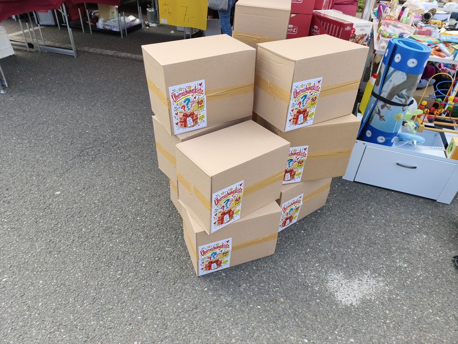 Gestapelte braune Kartons mit Aufkleber Ueberraschungskiste fuer 10 Euro, daneben Kinder- und Spielzeugware.