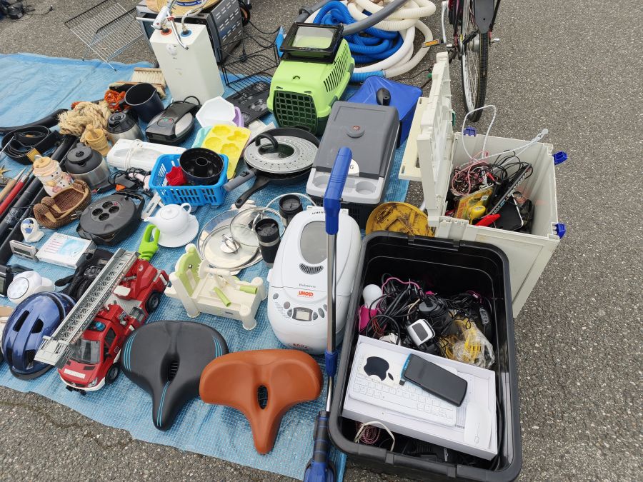 Draufsicht auf eine breite Warenauslage: Haushaltsgeraete, Kuechenutensilien, ein Feuerwehr-Spielzeugauto, Fahrradsaettel, ein Helm und Elektronik in einer Box liegen auf einer blauen Plane.