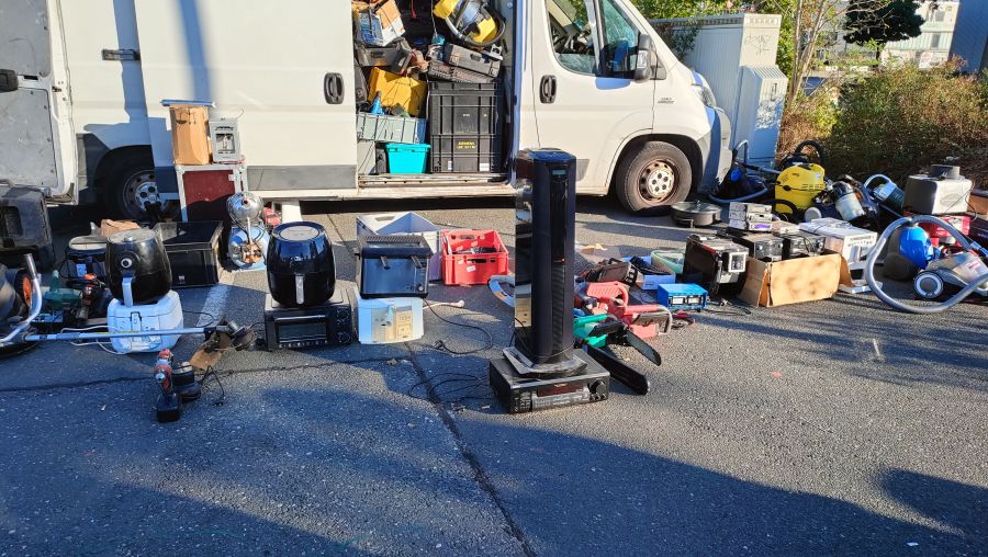 Flohmarktstand mit gebrauchten Elektrogeräten auf Asphalt vor einem weißen Transporter mit offenen Hecktüren: Heißluftfritteusen, HiFi-Lautsprechersäule, Staubsauger, Autoradios, Akkuschrauber und Werkzeug in Kisten.