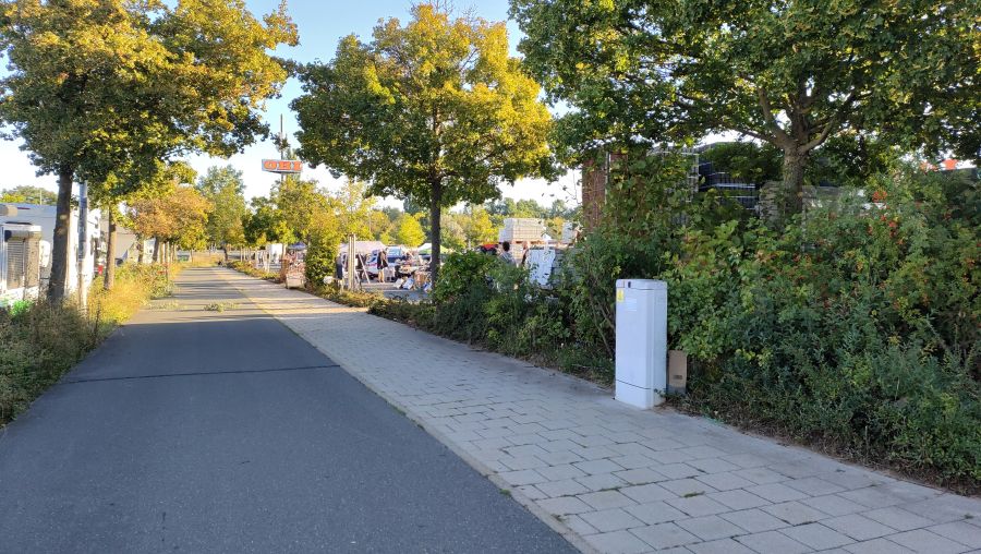 Zufahrt zum Flohmarkt auf dem OBI-Parkplatz Fürth: baumgesäumter Geh- und Radweg, im Hintergrund der OBI-Schriftzug auf dem Pylon und Verkaufsstände mit Pavillons.