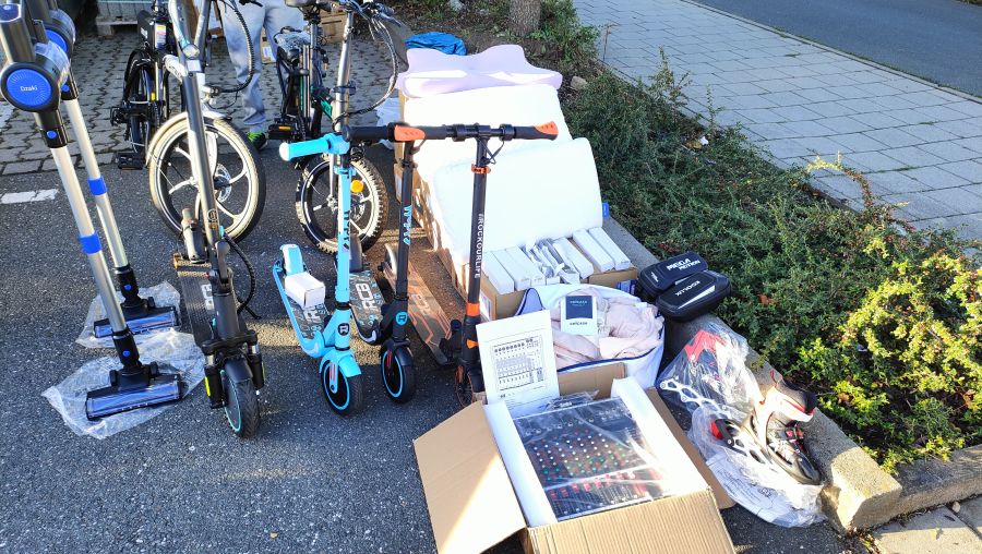 Verkaufsstand mit E-Scootern, akkubetriebenen Stielstaubsaugern, einem Kinderfahrrad, Inline-Skates, einem Mischpult und Matratzenauflagen auf dem Flohmarkt am OBI-Parkplatz Fürth.