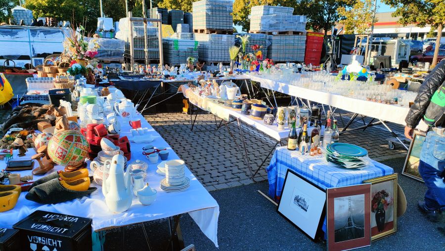 Mehrere Flohmarkttische mit Geschirr, Tassen, Teekannen, Trinkgläsern und Bierkrügen auf dem OBI-Parkplatz Fürth, im Hintergrund gestapelte Pflasterstein-Paletten.