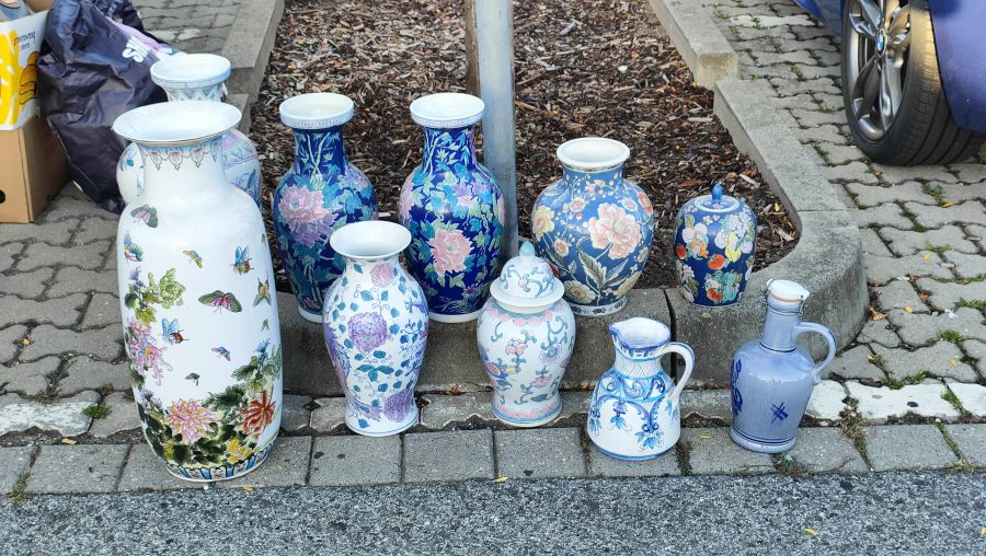 Reihe dekorativer Keramikvasen mit floralen Mustern, blau-weiße Ingwertöpfe und ein grauer Steinzeugkrug, am Bordstein aufgereiht auf dem Flohmarkt am OBI-Parkplatz Fürth.