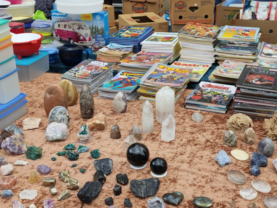 Auf einer Verkaufsdecke ausgebreitete Mineralien, Kristalle und Edelsteine, im Hintergrund gestapelte Comic-Hefte und ein Playmobil-Set.