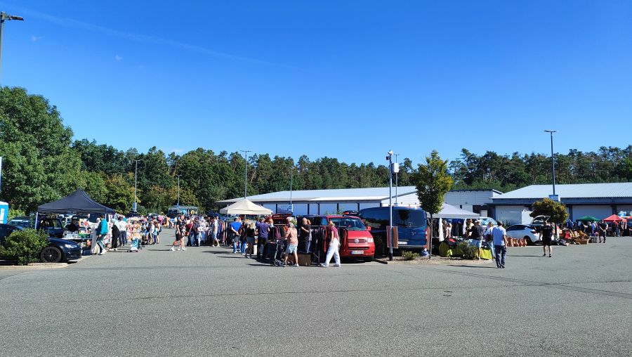 Weite Übersicht eines Flohmarkts auf einem großen Supermarkt-Parkplatz in Heßdorf mit einer langen Reihe Ständen, Besuchern und Verkäuferfahrzeugen vor Kiefernwald.