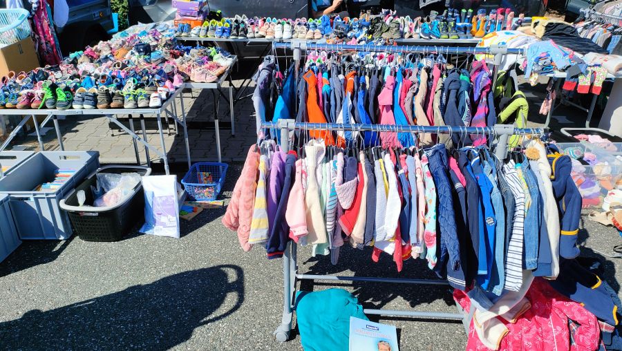 Volle Tische mit vielen Paar Kinderschuhen und zwei Kleiderständer mit Kinderjacken und Babykleidung auf einem Supermarkt-Flohmarkt.