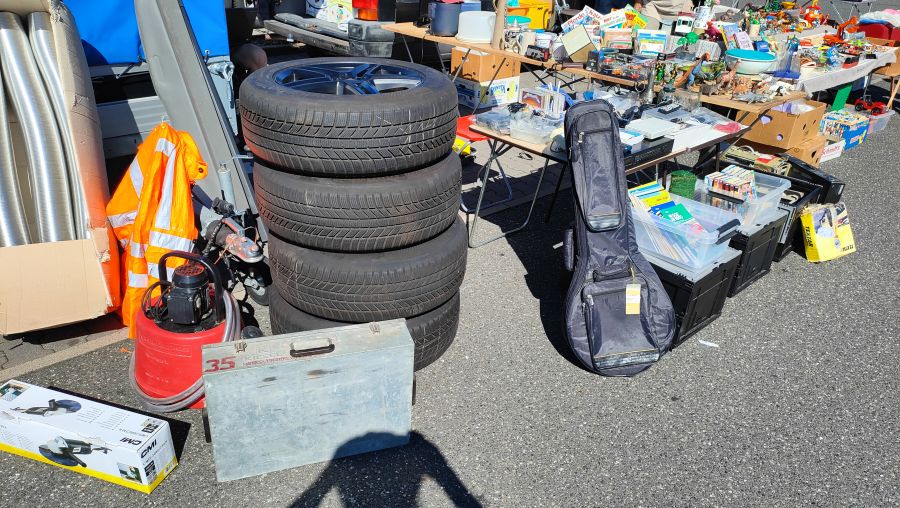 Vier gestapelte Alufelgen mit Winterreifen neben einer Tauchpumpe, einem Winkelschleifer-Karton und einer Gitarrentasche auf einem Flohmarkt.