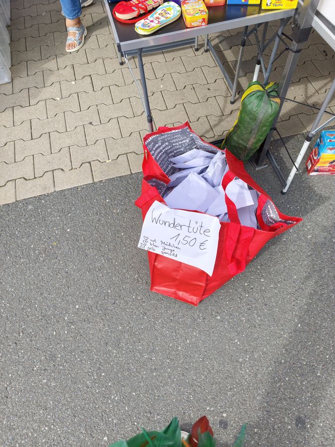 Rote Stofftasche voller in Papier gewickelter Ueberraschungspakete mit Schild Wundertuete 1,50 Euro, darueber ein Kindertisch mit Spielen.
