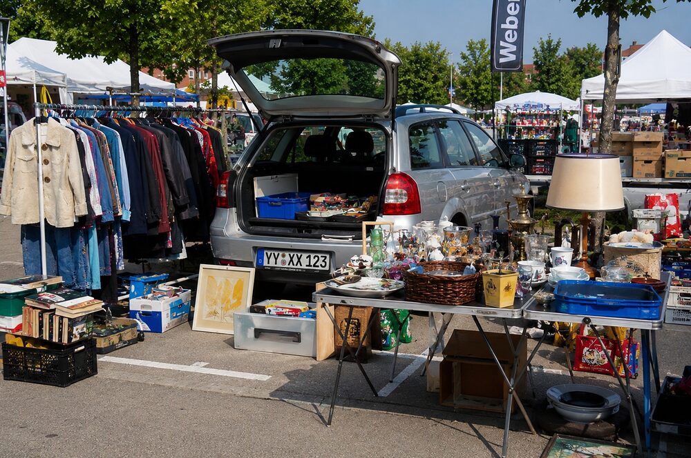 KI-generiertes Beispielbild eines Kofferraum-Flohmarkts: ein silberner Kombi mit offener Heckklappe, daneben eine Kleiderstange und ein Tisch voller Trödel, im Hintergrund Marktstände. Am Auto ist ein Kfz-Kennzeichen klar lesbar. (KI-generiertes Beispielbild)