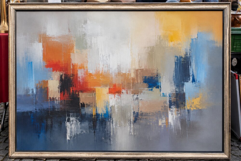 KI-generiertes Beispielbild: Ein einzelnes gerahmtes abstraktes Gemälde in Blau-, Orange- und Grautönen füllt fast das gesamte Bild. Vom Flohmarkt ist nur am Rand etwas zu sehen – eine rote Tischdecke, eine grüne Transportkiste, Kopfsteinpflaster. Keine Personen, keine Signatur. (KI-generiertes Beispielbild)