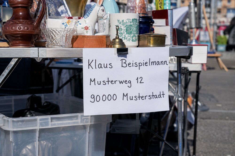 KI-generiertes Beispielbild eines Flohmarktstands: An der Kante eines Klapptischs mit Trödelware – Krüge, Keramik, eine Messingglocke – hängt ein handbeschriebenes DIN-A4-Blatt, darauf gut lesbar der Name „Klaus Beispielmann