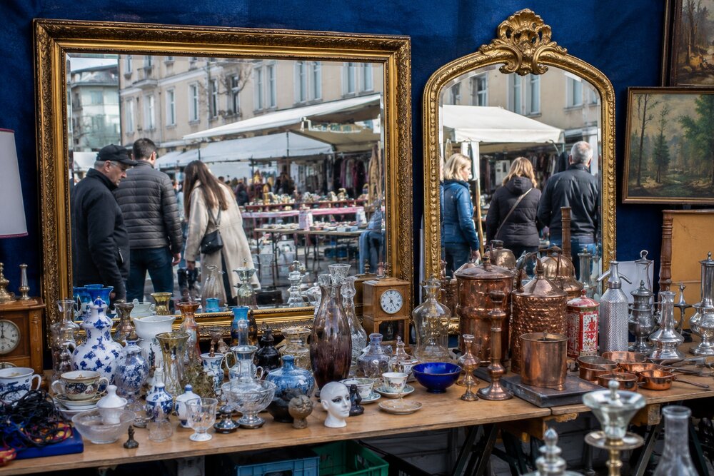 KI-generiertes Beispielbild eines Trödelstands mit zwei großen Standspiegeln in Goldrahmen vor blauem Tuch, davor ein Tisch voller Glas, Porzellan und Kupfergeschirr. In den Spiegeln spiegeln sich Marktbesucher und die Budenzeile gegenüber. (KI-generiertes Beispielbild)