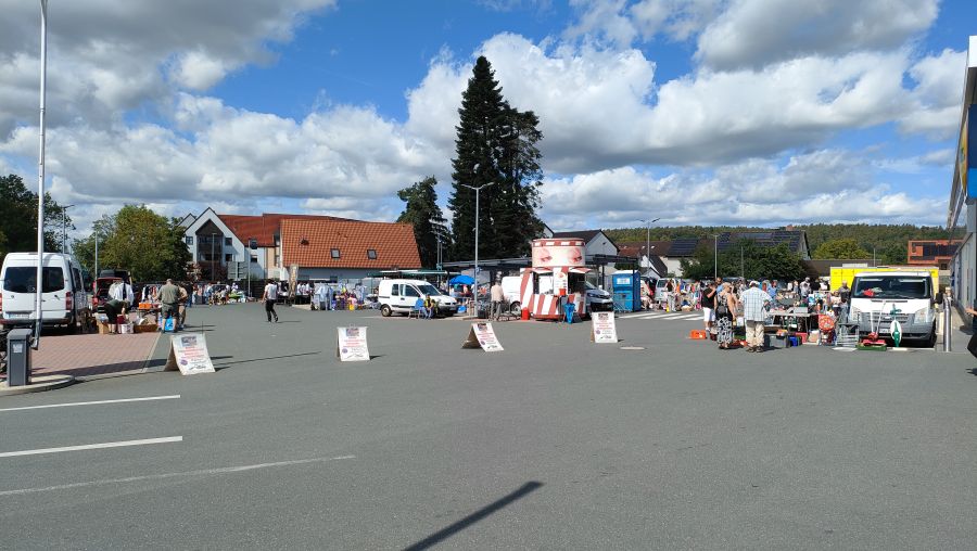 Überblick über den Flohmarkt auf dem LIDL-Parkplatz in Langenzenn: eine weite asphaltierte Fläche, an den Rändern Marktstände mit weißen Transportern, ein roter Imbisswagen und eine mobile Toilette. Auf dem freien Platz stehen „Parken verboten"-Aufsteller. Im Hintergrund eine große Kiefer, Wohnhäuser und das Marktgebäude. Sonniger Tag mit Wolken; Personen nur klein und aus der Entfernung.