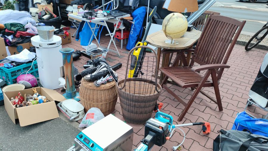 Bodenauslage auf dem Flohmarkt in Langenzenn: auf dem Pflaster stehen und liegen ein blauer Rollator, ein Weltkugel-Globus auf einem runden Holztisch, ein klappbarer Teak-Gartenstuhl, Inline-Skates, zwei geflochtene Körbe, ein Akku-Rasentrimmer, ein Kofferradio, ein Eimer und eine Kiste mit Garten-Deko-Figuren. Am linken Bildrand nur die Beine einer stehenden Person, kein Gesicht.