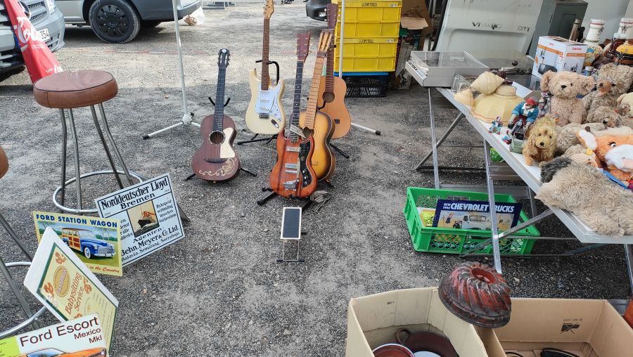 Mehrere gebrauchte Gitarren (Fender Stratocaster, Gibson-SG-artige E-Gitarre, Akustikgitarren) auf Ständern, umgeben von Retro-Blechschildern und Plüschtieren auf dem Flohmarkt.