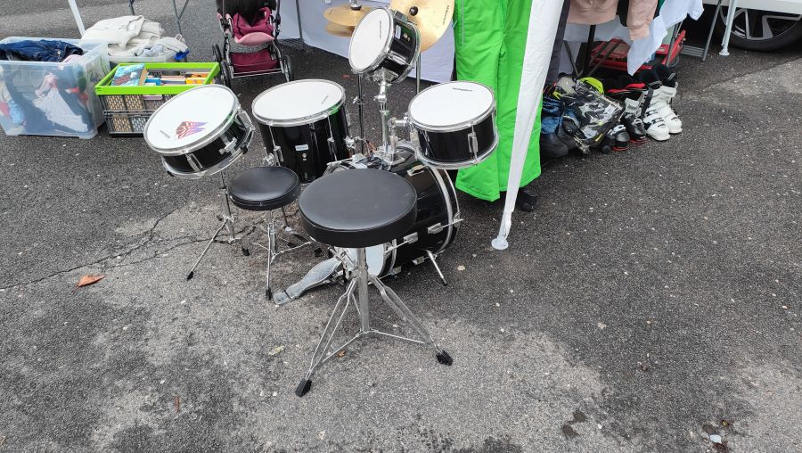 Komplettes schwarzes Millenium-Schlagzeug mit zwei Hockern auf dem Flohmarkt, im Hintergrund Kinderkleidung und Skischuhe.