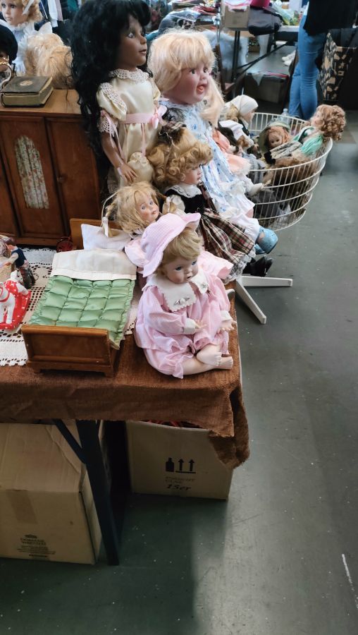 Sammlung antiker Porzellanpuppen mit Rüschenkleidern und ein Puppenbett auf einem Flohmarktstand.