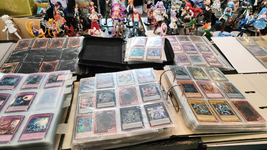 Ordner voller Yu-Gi-Oh!-Sammelkarten und dahinter eine Reihe bemalter Anime-Sammelfiguren (u.a. Sailor Moon) auf einem Flohmarktstand.