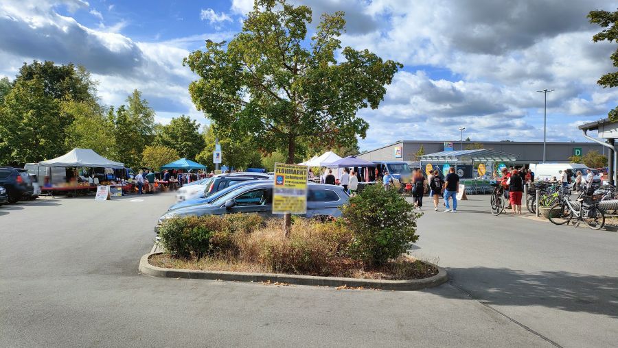 Blick ueber den Flohmarkt auf dem asphaltierten ALDI-Kundenparkplatz in Neunkirchen am Brand: eine Bauminsel im Vordergrund, dahinter parkende Autos und eine Reihe Staende mit Pavillons, rechts das ALDI-Gebaeude, bewoelkter Himmel.