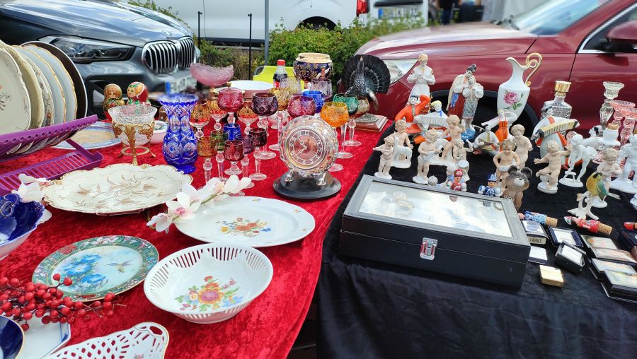 Antik- und Sammlerstand auf dem Flohmarkt in Neunkirchen am Brand: auf rotem Samt und schwarzem Tuch buntes boehmisches Bleikristall (Weinglaeser, Vasen), Bluetenteller aus Porzellan, eine Kristall-Tischuhr, Bisquit-Porzellanfiguren (Putten), ein Auerhahn-Praeparat und eine Vitrine mit Armbanduhren.