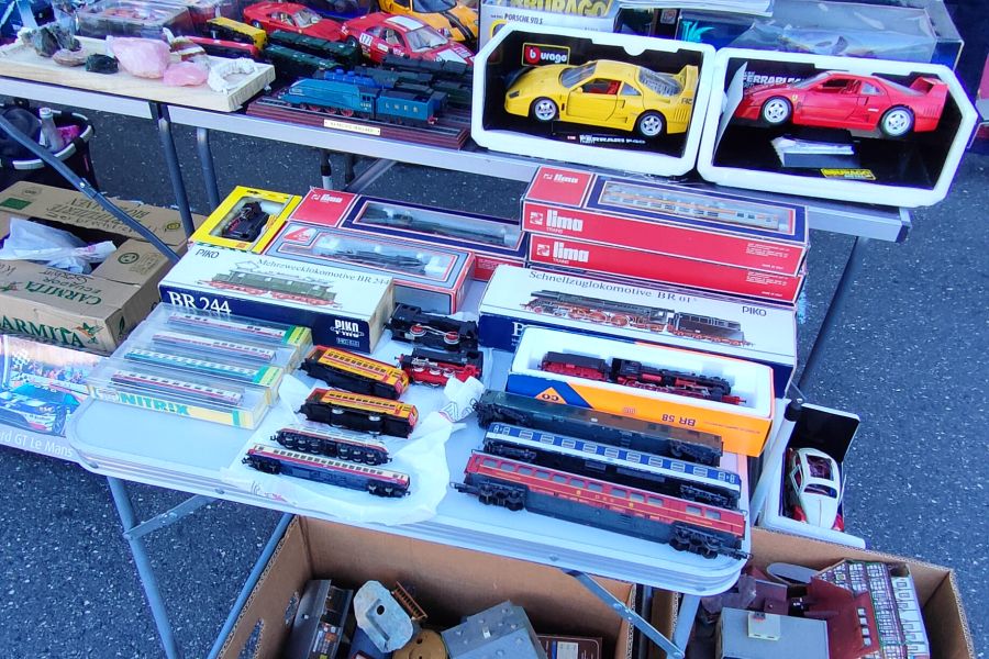 Querformat-Ausschnitt eines Flohmarkt-Sammlerstands: aufgereihte H0-Modelleisenbahn – Lokomotiven und Wagen von Lima, Piko und Roco, teils in Originalkartons (u. a. „BR 244") – auf einem weißen Klapptisch, darüber Modellautos von Bburago (gelber und roter Ferrari F40).