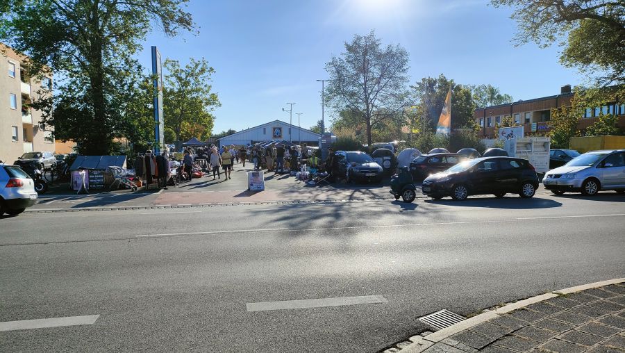 Flohmarkt auf einem ALDI-Gelände in Nürnberg-Eibach, von der Straße aus gesehen: eine Reihe Stände mit Kleiderständern und Pavillons, parkende Autos und ein Vespa-Roller, im Vordergrund eine breite Straße.