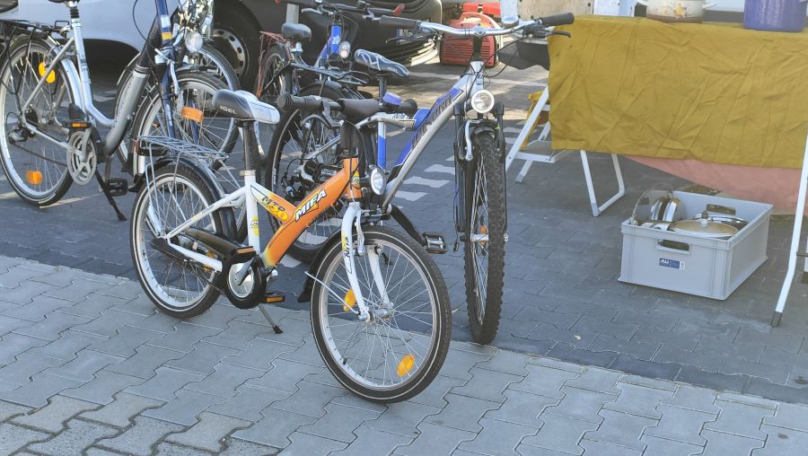Mehrere gebrauchte Fahrräder auf dem Asphalt eines Supermarkt-Flohmarkts: ein orange-weißes MIFA-Kinderrad im Vordergrund, dahinter Cityräder und ein Jugend-Mountainbike.