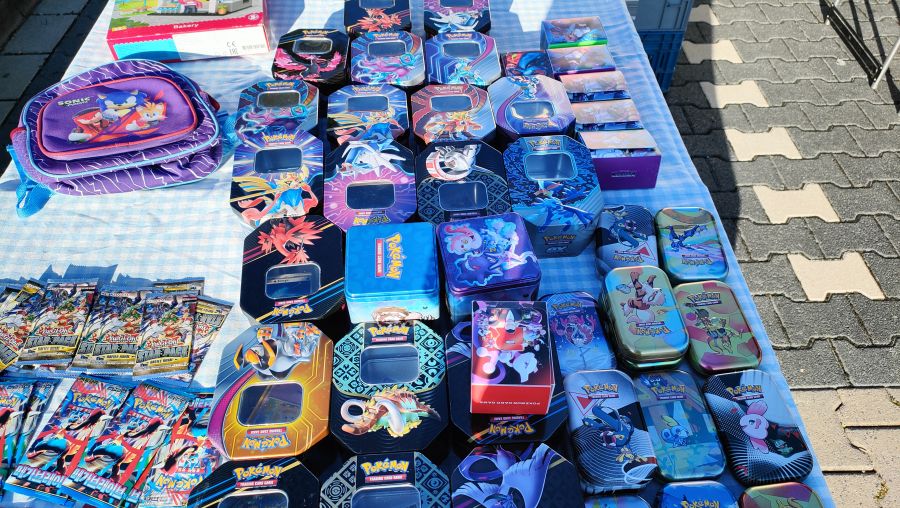 Tisch voller Pokémon-Sammelkarten-Blechdosen und Booster-Packungen sowie Yu-Gi-Oh!-Boostern auf einem Flohmarkt-Stand.