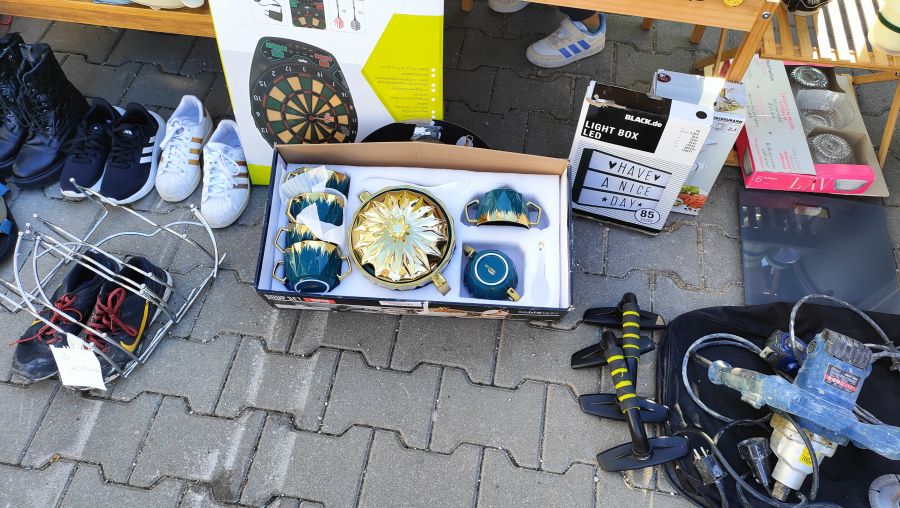 Bodenauslage mit Sneakern, Arbeitsstiefeln, einer elektronischen Dartscheibe, einem verpackten Tee-Service, einer LED-Lightbox und einem Farbsprühgerät auf einem Supermarkt-Flohmarkt.