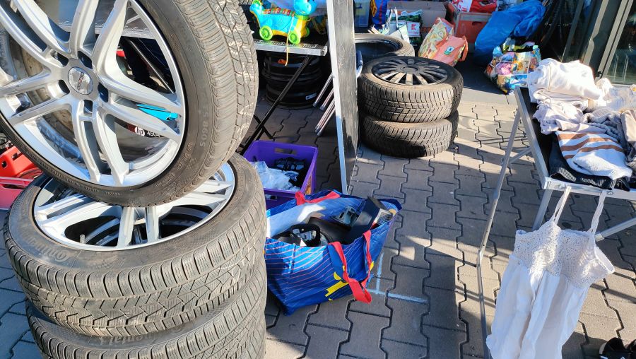 Gestapelte Alufelgen mit Winterreifen (DEZENT-Felgen, Continental-Reifen) auf dem Boden eines Supermarkt-Flohmarkts, daneben eine Kleiderstange mit einem weißen Häkelkleid.