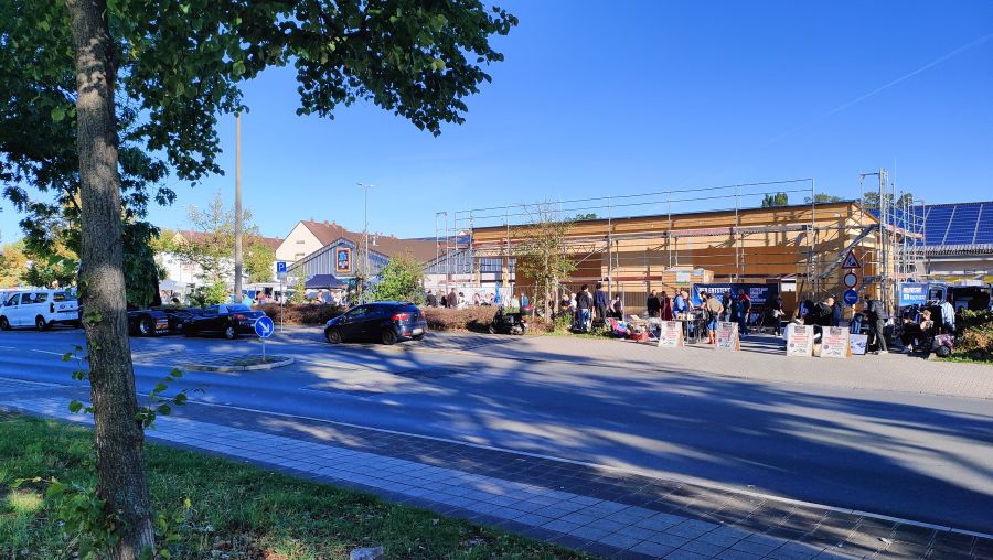 Flohmarkt auf einem ALDI-Parkplatz in Nürnberg, durch die Äste eines Straßenbaums gesehen, mit Ständen und Besuchern vor dem Supermarkt.