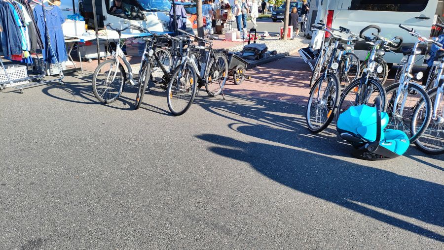 Reihe gebrauchter Trekking- und Cityräder auf einem Supermarkt-Parkplatz-Flohmarkt, im Vordergrund eine türkisfarbene Babyschale.
