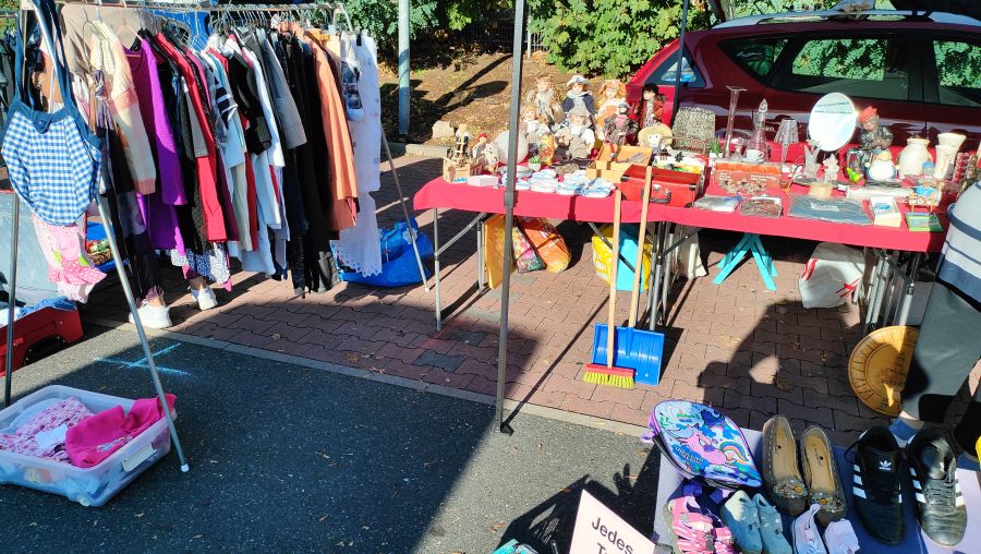 Flohmarktstand mit mehreren alten Porzellanpuppen und Matrjoschkas auf einem rot gedeckten Tisch, dahinter eine Kleiderstange mit Damenmode.