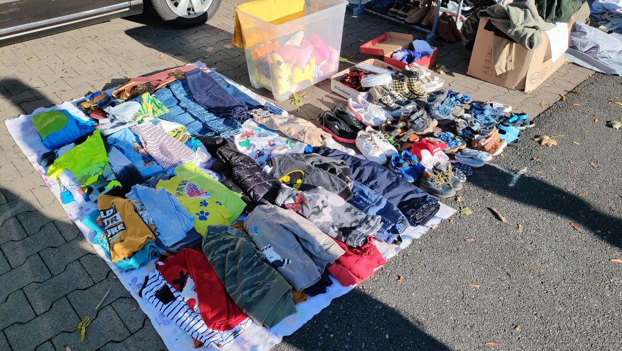 Auf einer Decke ausgebreitete gebrauchte Kinderkleidung und viele Paar Kinderschuhe, dazu eine Kiste Plüschtiere, auf einem Supermarkt-Flohmarkt.