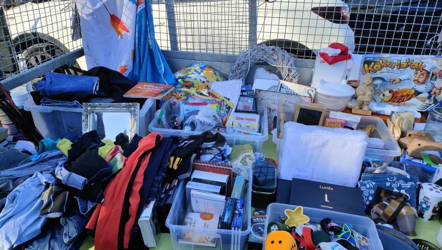 Auf einer grünen Decke am Boden ausgebreiteter Flohmarktstand mit hohen Stapeln gebrauchter Kleidung, durchsichtigen Sortierboxen voller Kleinkram und Bücher, dem Brettspiel Kakerlakak, einem silbernen Barock-Bilderrahmen, einer weißen Salatschleuder und einer kleinen Gartenzwerg-Figur; im Hintergrund der Gitteraufsatz eines Anhängers und geparkte Autos.