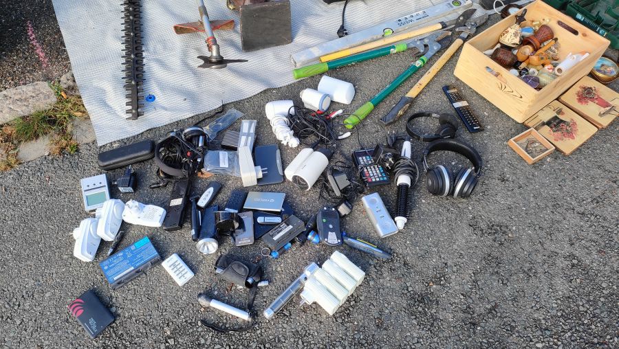 Zahlreiche gebrauchte Elektro-Kleinteile (Kopfhörer, Ladegeräte, Fernbedienungen, USB-Sticks) auf dem Asphalt ausgebreitet, daneben Gartenwerkzeug und eine Kiste mit bemalten Holz-Deko-Objekten.