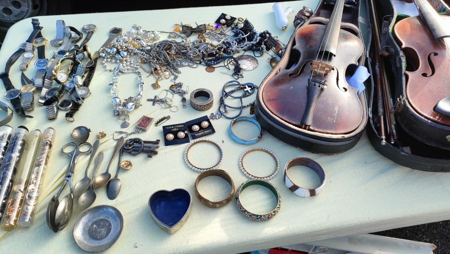 Schmuckstand mit vielen gebrauchten Armbanduhren, Halsketten, bunten Armreifen und Ohrringen sowie zwei alten Streichinstrumenten in Koffern.