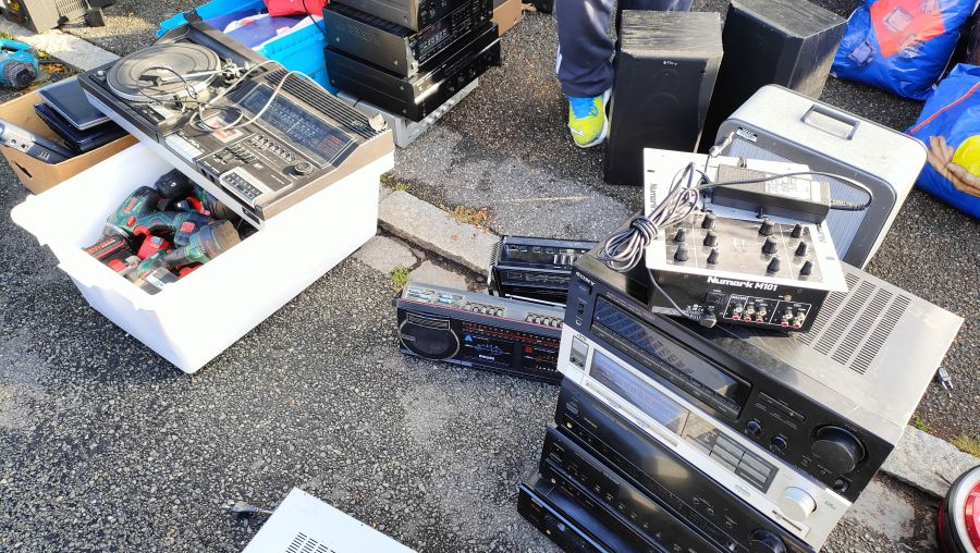 Sammlung gebrauchter Hi-Fi-Geräte (Musiktruhe mit Plattenspieler, Boomboxen, DJ-Mischpult Numark M101, Receiver) auf dem Asphalt, daneben eine Wanne mit Bosch-Akkuwerkzeugen.