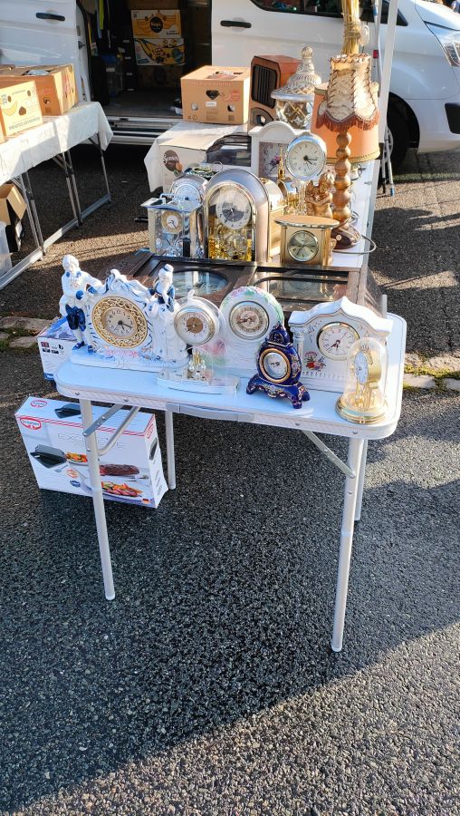 Sammlung gebrauchter Kaminuhren und Tischuhren, darunter eine Porzellan-Figurenuhr und eine Anniversary-Uhr unter Glasglocke, auf einem Flohmarktstand.