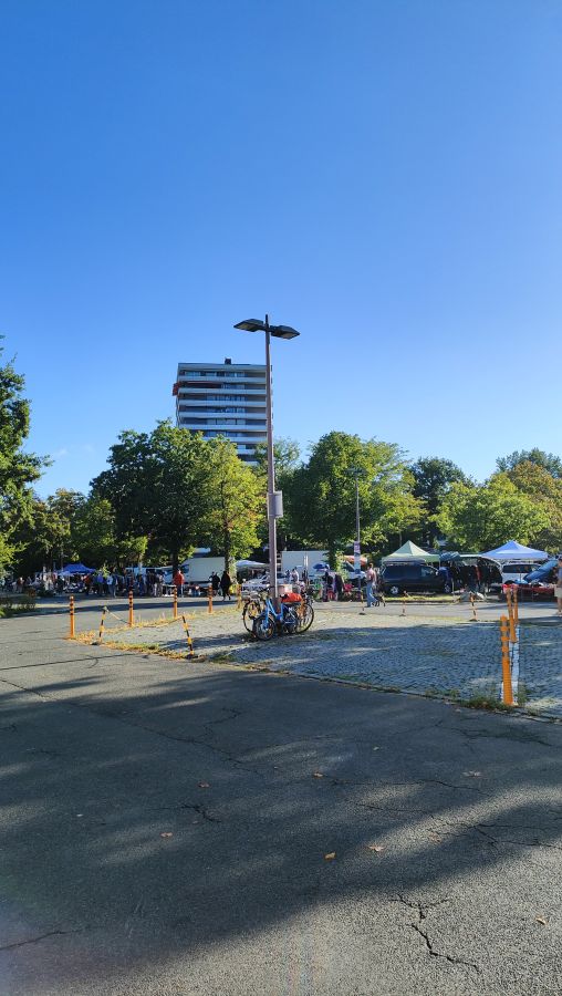 Hochformat einer großen leeren Parkplatzfläche mit Straßenlaterne und angeschlossenen Fahrrädern; im Mittelgrund die Ständereihe des Flohmarkts an der Meistersingerhalle Nürnberg mit Pavillons und Transportern vor einem Wohnhochhaus.