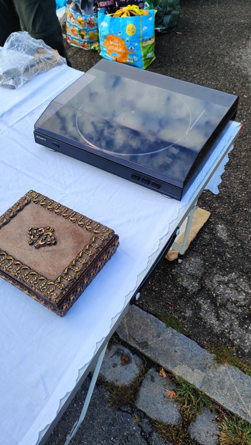 Gebrauchter Sony-Plattenspieler mit rauchglasfarbener Haube neben einer verzierten braun-goldenen Schmuckschatulle auf einem Flohmarkt-Verkaufstisch.