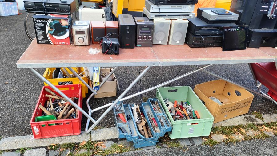 Flohmarktstand mit gebrauchter Hi-Fi-Elektronik (VHS-Recorder, JVC-Mikroanlage, Lautsprecher, Receiver, Plattenspieler) auf einem Tisch und mehreren Kisten voller Werkzeug darunter.